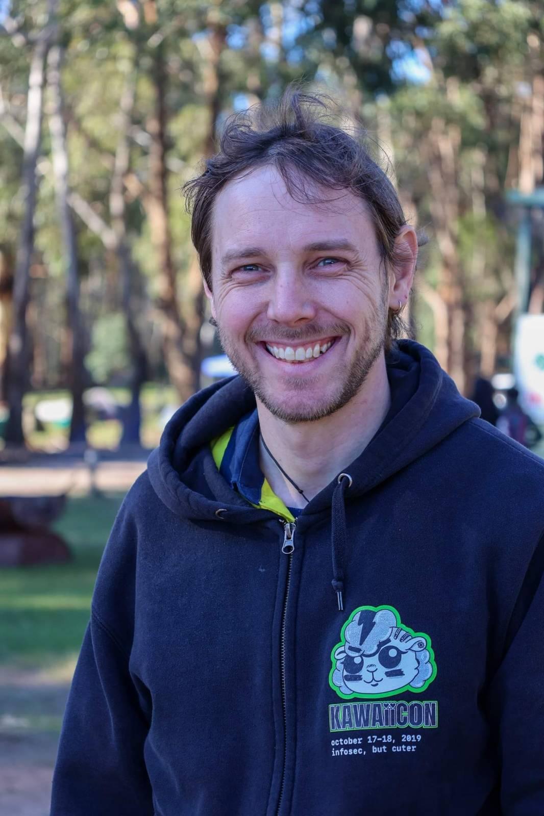 Matt, a masc-presenting person with a short beard, wind-blown hair,
and a wide grin, wearing a hoodie and staring at the camera. In the background
are some blurry but tall trees in dappled sunlight.